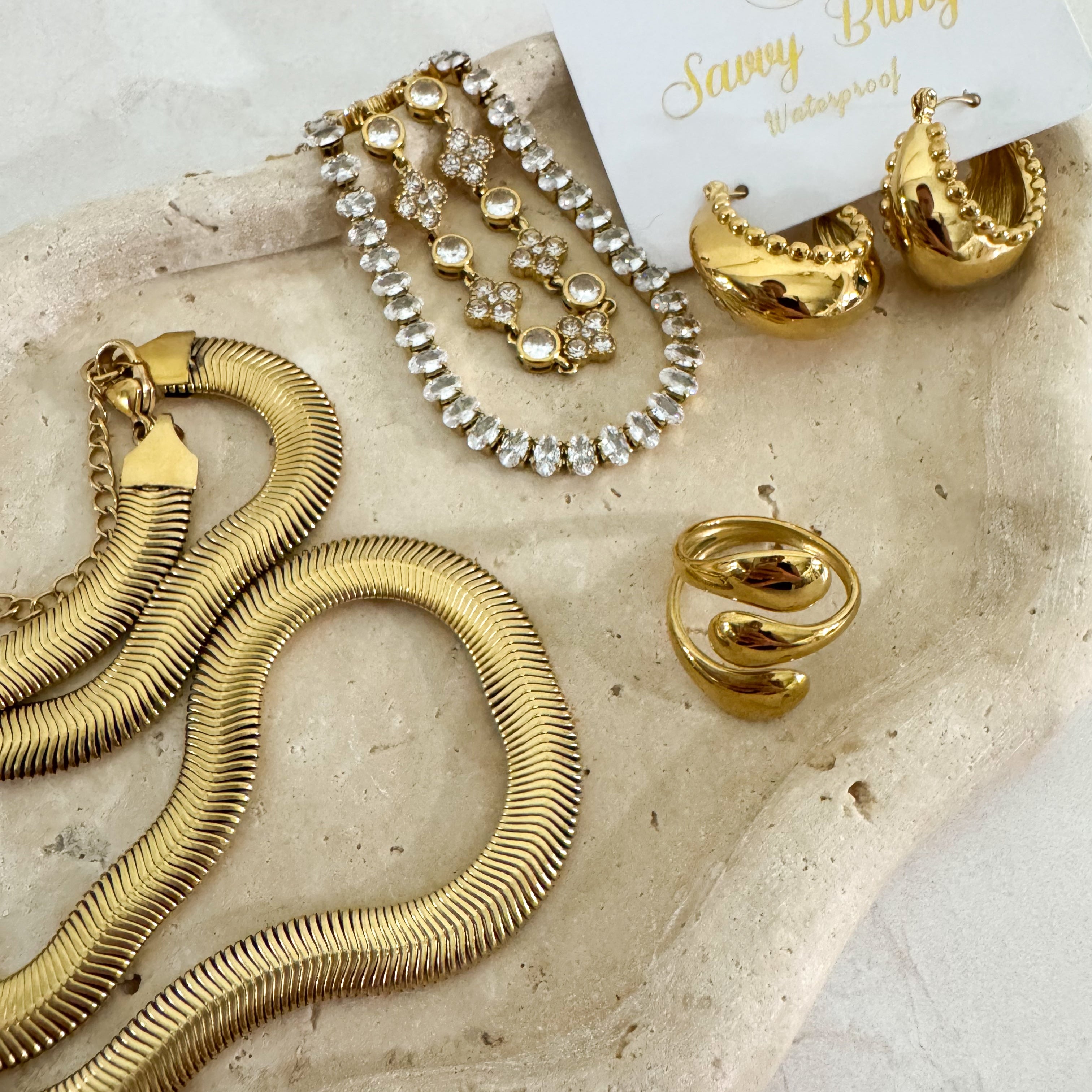 Waterproof Gold jewelry including necklace, bracelet, and hoop earrings on a stone surface with 'Savvy Bling' branding.