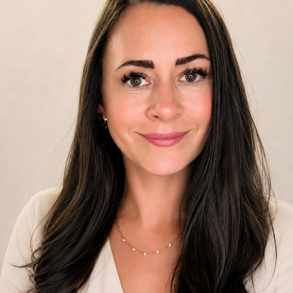 A woman with long, wavy dark brown hair is shown from the chest up against a plain, light beige background. She is wearing a cream-colored V-neck top and a delicate gold necklace with small green stones.
